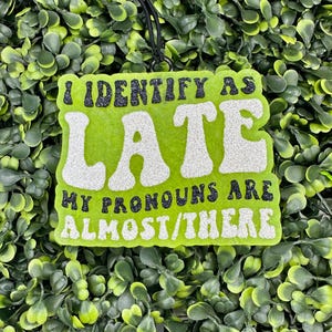 May include: Green and white glitter sign with the text "I identify as late, my pronouns are almost/there".