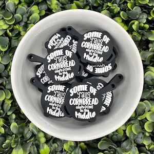 Puede incluir: Un bol lleno de adornos de cerámica negra y blanca con forma de sartenes de hierro fundido. Los adornos tienen el texto "Some of y'alls cornbread ain't done in the middle".