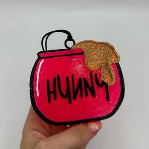 May include: A bright pink, rounded pot-shaped ornament with a black outline and a black hanging loop. The word HUNNY is written in black letters across the front. A textured, glittery gold substance drips over the top right edge of the pot.
