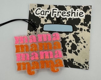 Könnte beinhalten: Ein glitzernder Auto-Lufterfrischer mit dem Wort mama, das viermal in abwechselnden Reihen in leuchtendem Pink und Orange wiederholt wird. Der Artikel ist an einer schwarzen Schnur befestigt und auf einer Karte mit schwarz-weißem Kuhmuster und dem Text Car Freshie abgebildet.