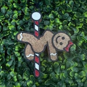 May include: A brown gingerbread man Christmas ornament with a black outline, glitter accents, and a red bow. The ornament hangs from a black string and is set against a backdrop of green foliage. The gingerbread man is holding onto a candy cane.