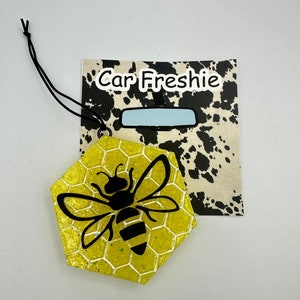 May include: Yellow hexagonal car freshener with a black bee silhouette on a honeycomb pattern. The freshener is attached to a black string and has a tag that says "Car Freshie".