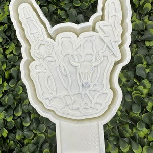 May include: White plastic mold of a skeleton hand making the rock and roll hand gesture. The hand has a lightning bolt, a skull, and a horseshoe on it.