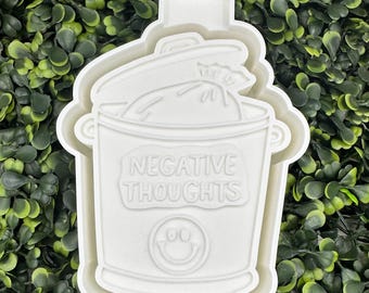 Może przedstawiać: Biała ceramiczna foremka do ciasteczek w kształcie garnka z pokrywką i workiem w środku. Garnek ma napisane na nim "NEGATIVE THOUGHTS" i uśmiechniętą buźkę na dole.