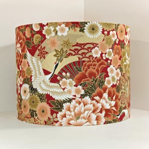 May include: Cylindrical lampshade with a Japanese design. The red fabric features a crane, flowers, and fan motifs in gold, white, and peach. The floral pattern includes peonies and other blossoms, creating a vibrant aesthetic.