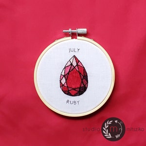 以下が含まれることがあります： 白い背景に「JULY」と「RUBY」の文字が刺繍された、赤いルビーの宝石を特徴とする刺繍のフープアートです。