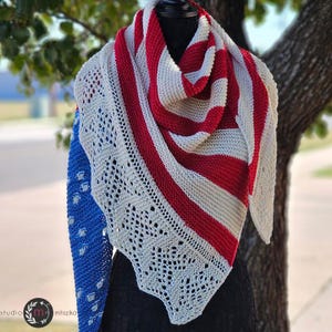 May include: A white and red striped knitted shawl with a crocheted lace border. The shawl is draped over a mannequin and is displayed against a blurred background of green trees.
