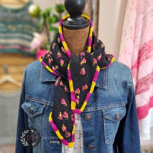 May include: A dark gray knitted scarf with a floral pattern in pink and yellow. The scarf has a yellow and pink border and is displayed on a mannequin wearing a denim jacket. The scarf is a handmade accessory.
