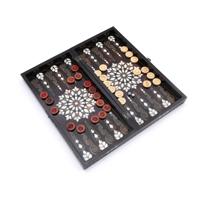 Peut inclure: Un jeu de backgammon en bois noir avec un motif en nacre. Le plateau est ouvert et montre les pions, qui sont rouges et beiges. Il y a deux d&eacute;s sur le plateau.