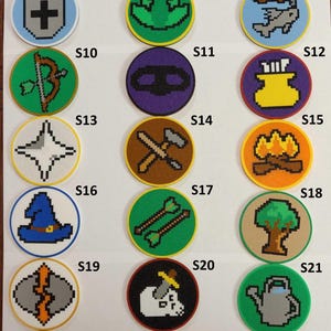 Pode incluir: Uma coleção de 26 patches circulares com designs pixelizados. Cada patch apresenta um ícone diferente, incluindo uma espada, um coração, uma picareta, uma mão, uma figura correndo, uma bigorna, uma cruz, um sapo, um peixe, arco e flechas, uma máscara, um tinteiro, uma estrela, um martelo e uma picareta, uma fogueira, um chapéu de mago, flechas, uma árvore, um raio, um cão, um regador, uma pegada, uma harpa, uma bússola e uma explosão de estrelas.