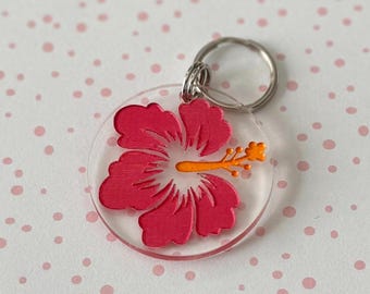 May include: A pink hibiscus flower keychain with a yellow center and a silver key ring. The flower is on a clear acrylic background.