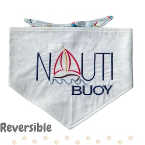 May include: White dog bandana with embroidered text "NAUTI BUOY" and a sailboat design in red, yellow, and brown. The bandana has a blue and white patterned collar. The word "Reversible" is printed at the bottom.