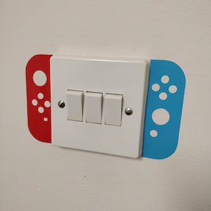 May include: A white light switch with three buttons, decorated with a red and blue Nintendo Switch controller design.