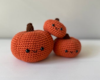 Puede incluir: Tres calabazas de crochet con caras sonrientes. Las calabazas son de color naranja y tienen ojos y bocas negros. La calabaza más grande está en la parte delantera y la más pequeña en la parte trasera.