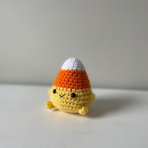 Könnte beinhalten: Ein gehäkeltes Plüschspielzeug in Form eines Maisbonbons mit einem Farbschema aus Weiß, Orange und Gelb. Das Spielzeug hat ein süßes, cartoonartiges Gesicht mit schwarzen Augen und einem kleinen Lächeln.