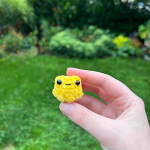 May include: A small, yellow crocheted plush toy shaped like a frog with black eyes. The toy is being held in a hand.