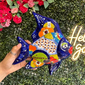 May include: A hand-painted ceramic fish plate in vibrant colors. The fish features a deep blue base with colorful floral and geometric patterns in orange, yellow, green, and white. The plate has a glossy finish and is held against a green backdrop.