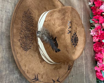 May include: Brown suede cowboy hat with decorative feather and white band. The hat features etched designs, including a feather and geometric patterns. A stylish accessory for a western-themed look.