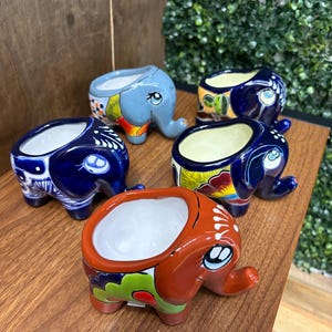 May include: Five ceramic elephant-shaped planters featuring hand-painted designs in various colors including blue, light blue, and orange. Each planter has a hollow center, a trunk, and decorative patterns on the sides with white interiors.