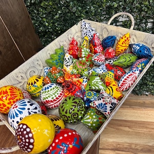 May include: A variety of colorful, hand-painted ceramic ornaments in a distressed white tray. The ornaments are various shapes, including spheres and teardrops, with intricate patterns in red, yellow, blue, and green. The tray has a handle.