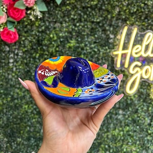 May include: A vibrant, hand-painted ceramic sombrero ashtray. The ashtray features a deep blue crown and a colorful brim with orange, green, and white floral designs. The ashtray is held in a hand against a green background with a neon sign.