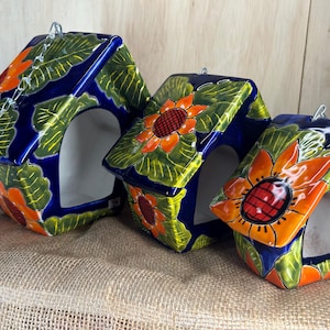 May include: Three ceramic birdhouses painted in a vibrant blue, green, and orange floral pattern. Each house has a small opening for birds to enter and a chain for hanging.