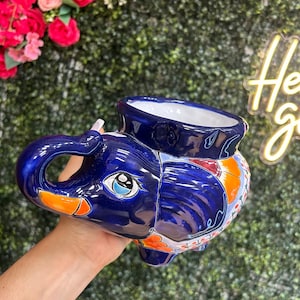 May include: A blue ceramic elephant-shaped planter with orange and white floral accents. The planter has a large opening on top and is held by a hand. The elephant has a detailed trunk, ears, and eyes.