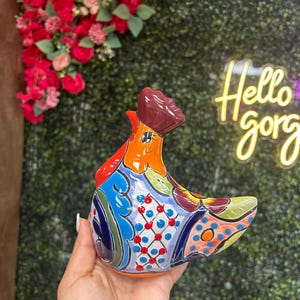 May include: A colorful ceramic rooster figurine with a detailed, hand-painted design. The rooster features vibrant hues of blue, orange, red, and green, with intricate patterns. The text "Hello gorg" is visible in the background.