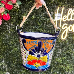 May include: A hand-painted ceramic hanging planter with a vibrant floral design in blue, orange, red, and yellow. The planter has a rope handle and metal rings for hanging. The background is a green hedge.