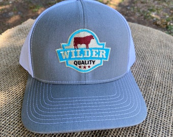 May include: Gray and white trucker hat with a blue and white logo that says "Wilder Quality" and features a red silhouette of a cow.