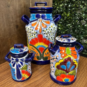 May include: Three hand-painted ceramic milk jugs with lids. The jugs are decorated with vibrant floral and geometric patterns in blue, orange, red, and green. Each jug has a handle and a small handle on the lid.