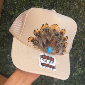 May include: A beige trucker hat with a mesh back and a brown and yellow feather accent. The hat has a label that reads "Otto World Famous Foam Front OSFM Trucker Durable Snap Closure".