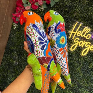 May include: Two ceramic parrot figurines featuring vibrant hand-painted Talavera-style patterns. One parrot is primarily orange with blue and green accents, while the other is green with blue and red floral motifs. Both have curved beaks.