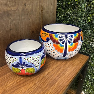 May include: Two decorative ceramic planters with a vibrant, hand-painted design. The planters feature a white interior, a blue rim, and colorful floral and geometric patterns in orange, blue, green, and red. They are placed on a wooden shelf.