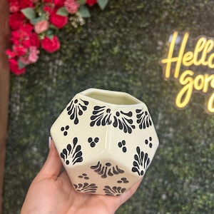 May include: Cream-colored, geometric ceramic vase with black floral patterns and small dots. The vase is held in a hand, with a blurred background of flowers and a neon sign that says "Hello gorgeous".