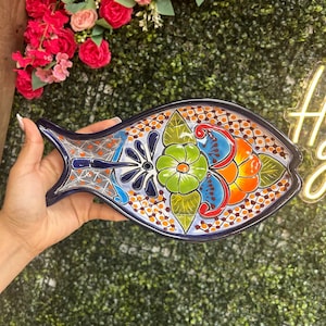 May include: A hand-painted, ceramic fish-shaped serving dish. The dish features a vibrant floral design in green, orange, and blue, with a dark blue border. The background includes a green hedge and pink flowers.