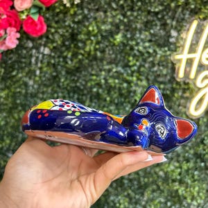 May include: A ceramic cat figurine in a reclining pose. The cat is primarily dark blue with red, yellow, and white accents. The figurine has a glossy finish and is held in a hand.