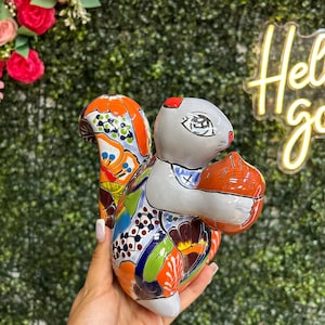 May include: A ceramic squirrel figurine with a gray body, holding a brown nut. The squirrel's tail and body are decorated with colorful patterns in orange, blue, green, and white. The figurine is held in a hand.
