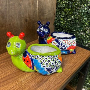 May include: Three colorful ceramic planters. One is shaped like a green snail with red accents, another is a blue creature, and the third is a blue and white rectangular pot with floral designs. The planters are on a wooden surface.