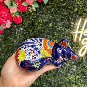 May include: A hand-painted ceramic fox figurine in a sleeping pose. The fox is primarily blue with colorful accents of orange, yellow, red, and white. The figurine is held in a hand against a backdrop of green foliage and red flowers.