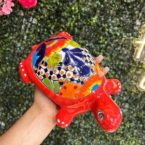 May include: A vibrant, hand-painted ceramic turtle figurine. The turtle is primarily red with a colorful shell featuring floral and geometric patterns in orange, blue, white, and green. The figurine has a glossy finish and is held in a hand.