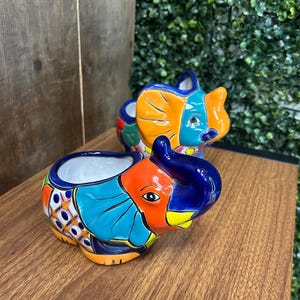 May include: Two colorful ceramic elephant planters. The elephants are painted with bright orange, blue, yellow, and green patterns. The planters have a white interior and are sitting on a wooden surface. The elephant planters are a decorative item.