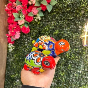 May include: Two colorful ceramic turtle figurines. The turtles have red, orange, yellow, blue, and green patterns. The larger turtle is approximately 4 inches long, and the smaller turtle is about 3 inches long. The background features a green wall and red flowers.