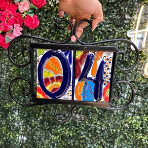 May include: Black wrought iron house number holder with colorful ceramic tiles displaying the number "04". The tiles feature a vibrant floral design with blue, orange, and red hues. The holder is held up against a green hedge.