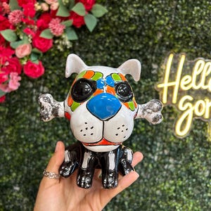 May include: A ceramic dog figurine with a Day of the Dead design. The dog has a white head with colorful accents, black eyes, and a blue nose. It holds a bone in its mouth and has black and white markings.