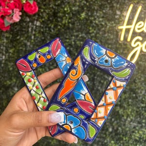 May include: Two hand-painted, blue ceramic frames with colorful floral and geometric designs. The frames feature orange, green, red, and yellow accents. The frames are held in a hand, with a background of greenery and flowers.