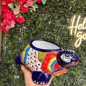 May include: A ceramic turtle-shaped planter with a vibrant, hand-painted design. The planter features a blue base and head, with colorful patterns of orange, yellow, and blue on its shell. The interior is white, and the overall design is whimsical and decorative.