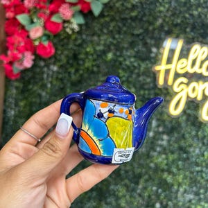 May include: A small, hand-painted ceramic teapot in shades of blue, yellow, and orange. The teapot features a curved handle and spout, with a lid on top. The words "HECHO EN MEXICO" are printed on a small label.