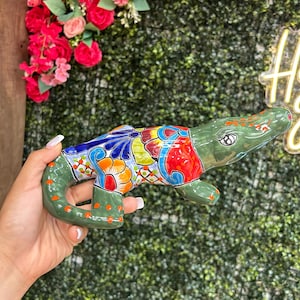 May include: A ceramic alligator figurine with a green body and colorful, intricate patterns on its back. The design includes blue, orange, red, and yellow floral motifs. The alligator is held in a hand against a backdrop of greenery and flowers.
