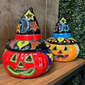 May include: Two ceramic Halloween pumpkin jars with witch hat lids. The pumpkins are red and orange with carved faces and colorful floral designs. The black witch hats have star and moon cutouts.